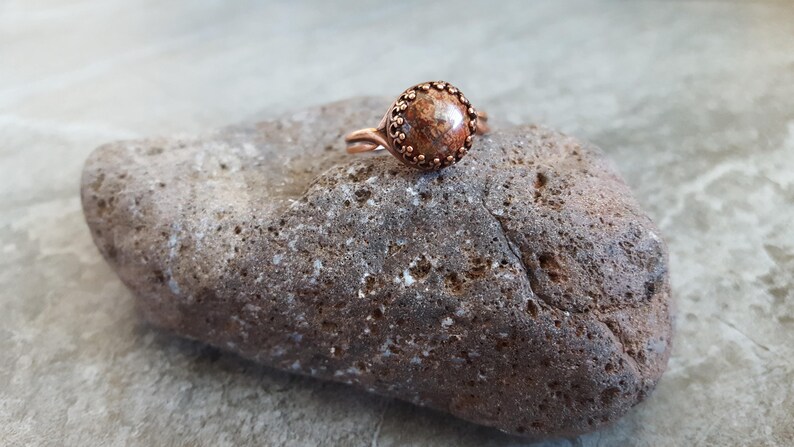 May include: A copper ring with a brown and orange stone set in a copper bezel. The ring is sitting on a rough, gray rock.