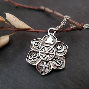 May include: Silver-toned flower pendant necklace featuring symbols of various religions. The pendant has a central sun-like design surrounded by symbols including a cross, peace sign, and star and crescent. The necklace is displayed on a dark surface.