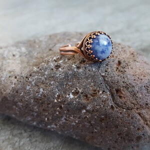 May include: A copper ring with a blue and white stone set in a decorative copper bezel. The ring is resting on a rough, gray rock.
