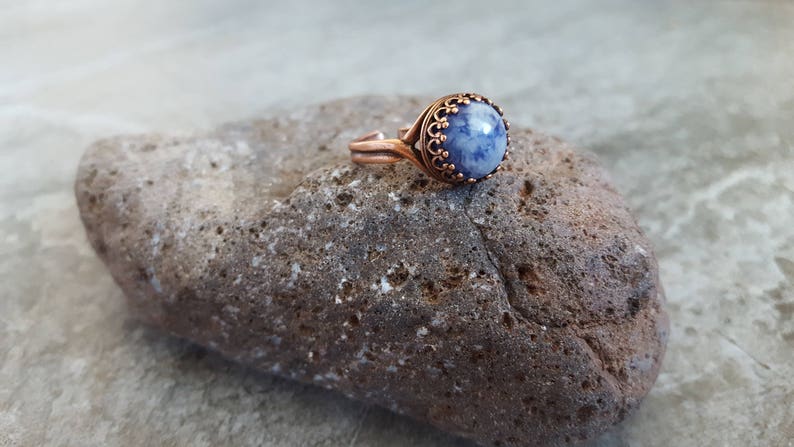 May include: A copper ring with a blue and white stone set in a decorative copper bezel. The ring is resting on a rough, gray rock.