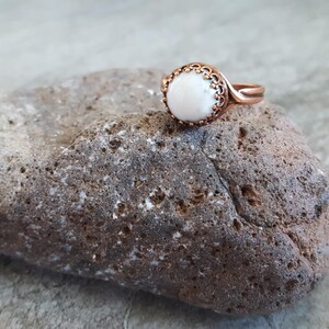 May include: A copper ring with a white stone set in a decorative bezel. The ring is resting on a brown rock.