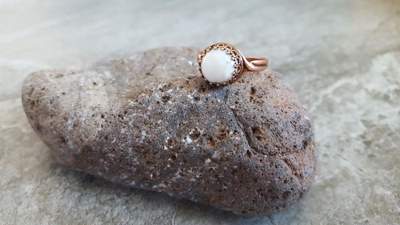 May include: A copper ring with a white stone set in a decorative bezel. The ring is resting on a brown rock.