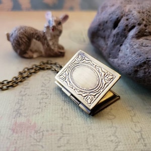 May include: A gold-toned book-shaped locket necklace with an ornate design and a blank oval space in the center. The locket is open and the chain is visible.