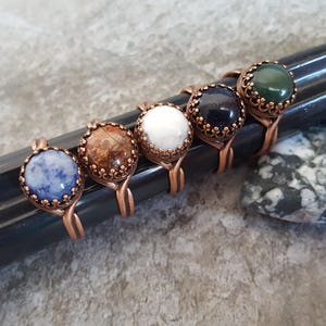 May include: Six copper wire rings with different colored gemstone cabochons. The stones are blue, brown, white, black, green, and blue.