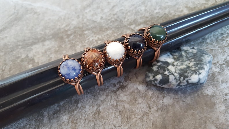 May include: Six copper wire rings with different colored gemstone cabochons. The stones are blue, brown, white, black, green, and blue.