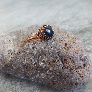 May include: A copper ring with a dark blue gemstone set in a decorative bezel. The ring is sitting on a rough, gray rock.
