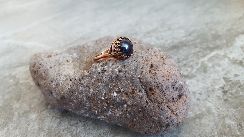 May include: A copper ring with a dark blue gemstone set in a decorative bezel. The ring is sitting on a rough, gray rock.