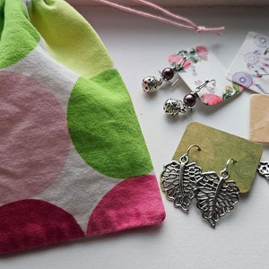 May include: A collection of earrings displayed on small cards, alongside a drawstring bag. The earrings feature various designs, including leaf shapes, floral arrangements, and beaded styles. The bag is white with large pink and green polka dots.