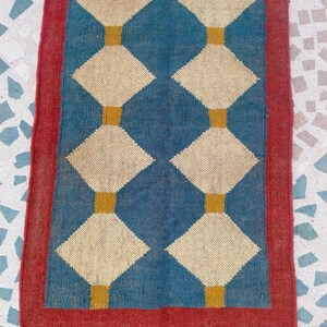 May include: A rectangular rug with a blue centre featuring a diamond pattern in cream and gold. The rug has a red border and a fringe at the bottom. The rug is made of woven material.
