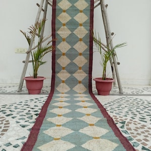 May include: A long rectangular table runner featuring a repeating diamond pattern in light blue and cream, accented with small mustard yellow squares at each intersection. The runner has a dark maroon border and fringed ends on both sides.