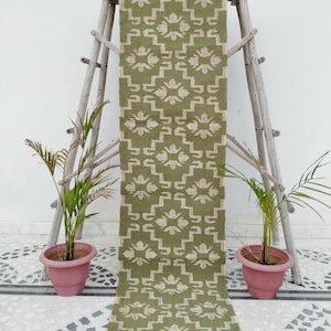 May include: A long, rectangular table runner in olive green featuring a repeating cream-coloured geometric pattern of stepped lines and floral-like motifs. The runner has a textured, woven appearance with short, frayed fringe edges at the bottom.