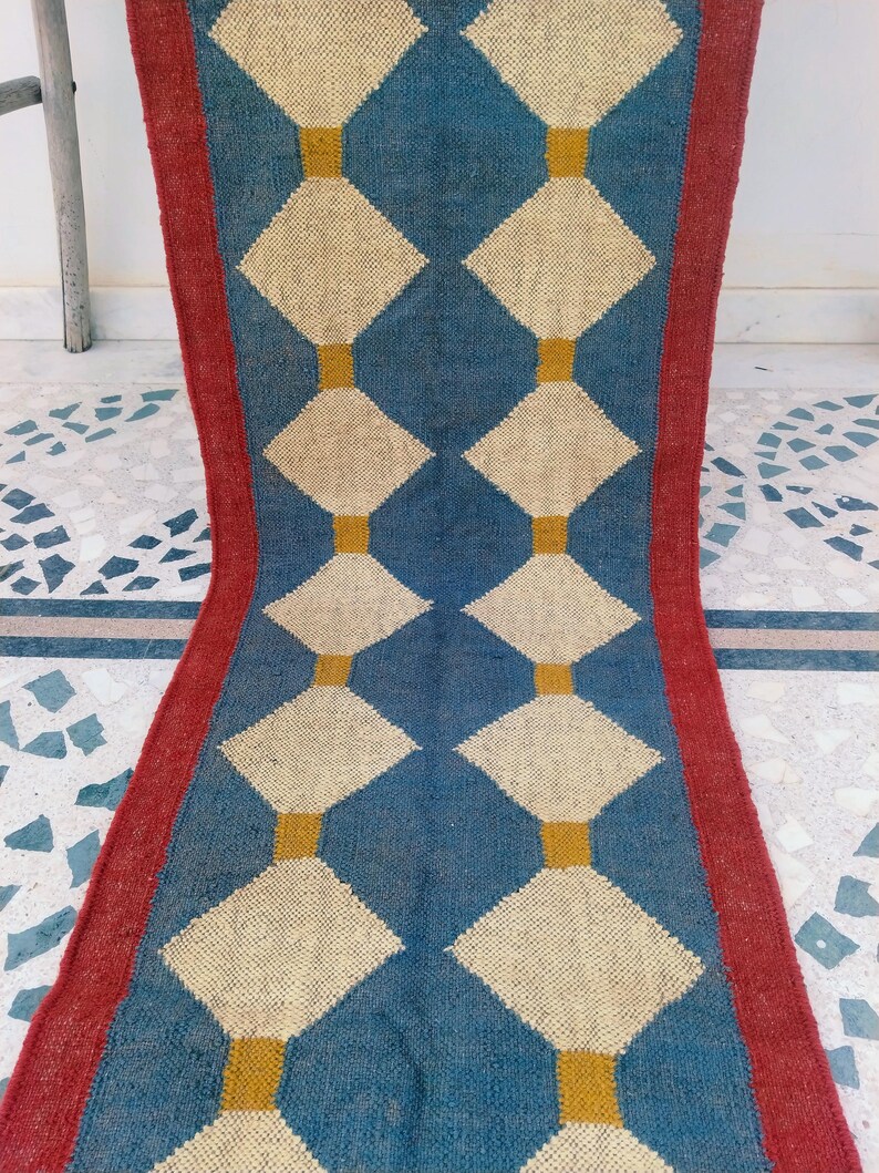 May include: A blue and cream-colored runner rug with a repeating diamond pattern. The diamonds are cream-colored and outlined in yellow, set against a blue background. The rug has a red border.