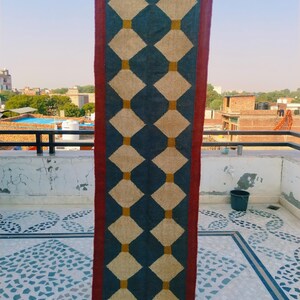 May include: A rectangular rug with a geometric pattern. The rug features a dark teal background with a repeating pattern of cream-colored diamonds connected by gold squares. A burgundy border frames the design.