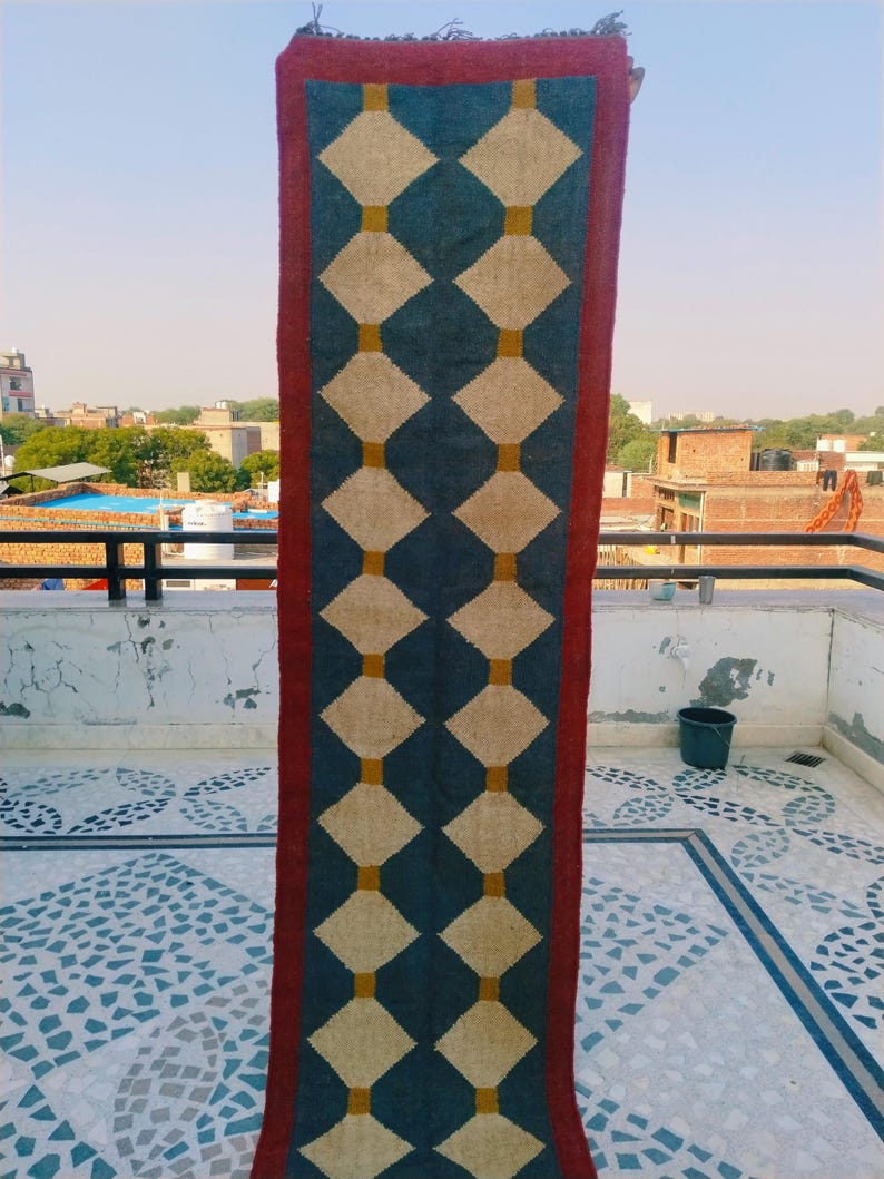 May include: A rectangular rug with a geometric pattern. The rug features a dark teal background with a repeating pattern of cream-colored diamonds connected by gold squares. A burgundy border frames the design.