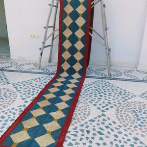 May include: A long, rectangular rug with a repeating diamond pattern. The rug features a teal blue background with cream-colored diamonds and a red border. The rug is displayed on a wooden ladder.