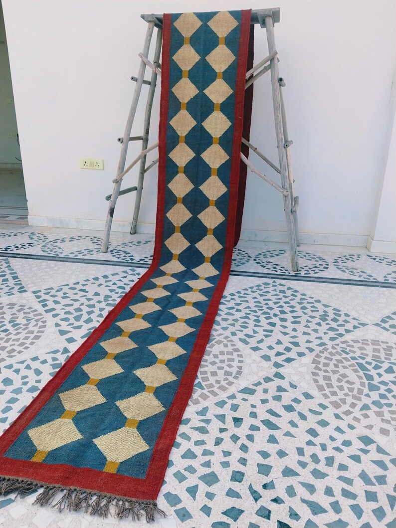 May include: A long, rectangular rug with a repeating diamond pattern. The rug features a teal blue background with cream-colored diamonds and a red border. The rug is displayed on a wooden ladder.