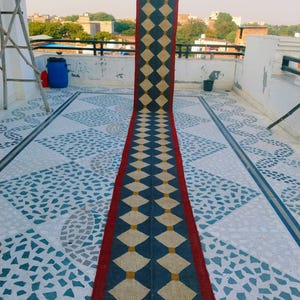 May include: A long, rectangular rug with a repeating diamond pattern. The rug features a dark blue and cream diamond design with a red border and fringe. The rug is displayed outdoors on a tiled surface.