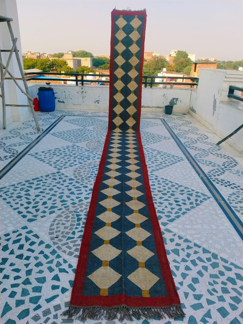 May include: A long, rectangular rug with a repeating diamond pattern. The rug features a dark blue and cream diamond design with a red border and fringe. The rug is displayed outdoors on a tiled surface.