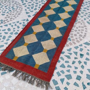 May include: A rectangular runner rug with a blue diamond pattern and a red border. The diamonds are beige and connected by yellow lines. The rug has a fringe on one end.
