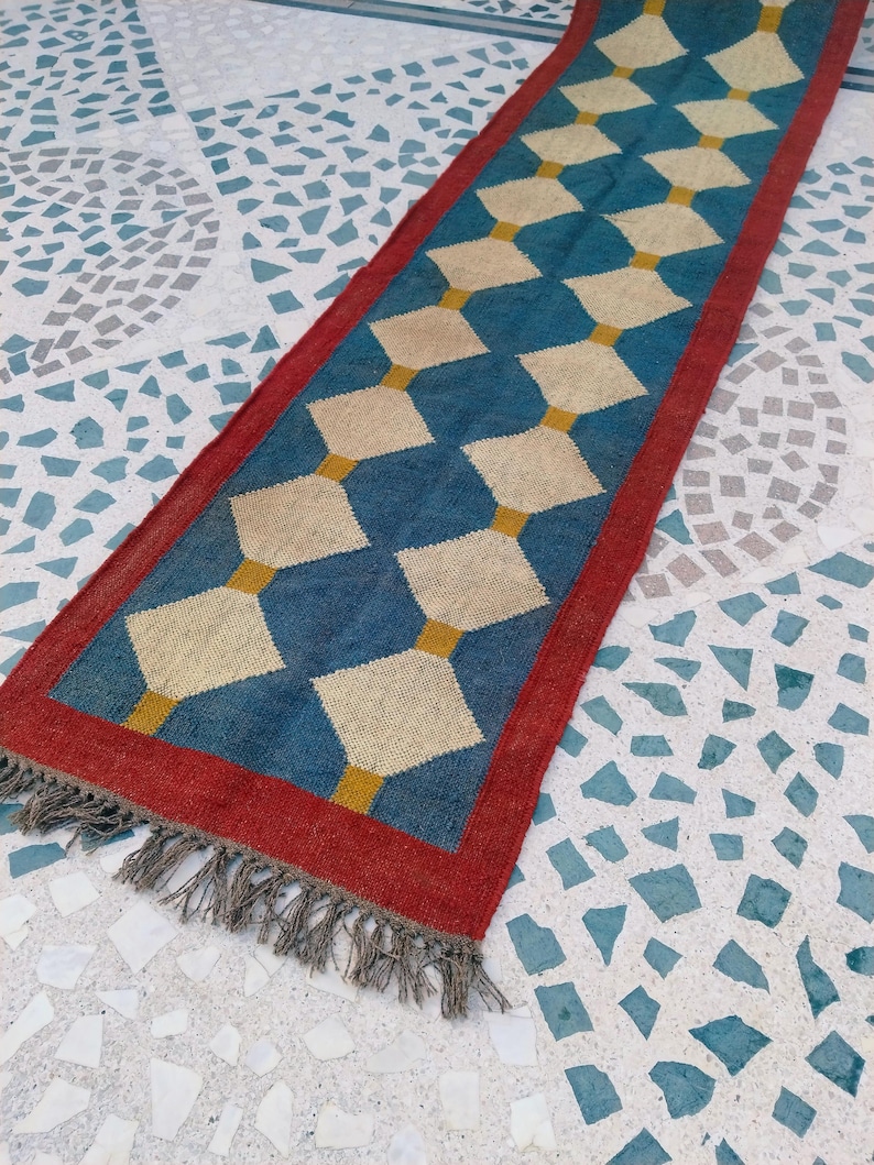 May include: A rectangular runner rug with a blue diamond pattern and a red border. The diamonds are beige and connected by yellow lines. The rug has a fringe on one end.