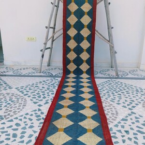 May include: A long, rectangular rug with a repeating diamond pattern. The rug features a blue background with cream-colored diamonds and yellow accents. A red border frames the design, and the rug has a fringed edge. The rug is displayed on a patterned floor.