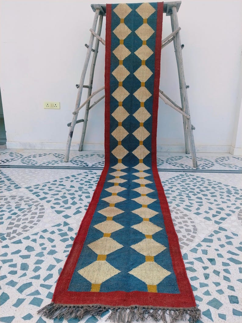 May include: A long, rectangular rug with a repeating diamond pattern. The rug features a blue background with cream-colored diamonds and yellow accents. A red border frames the design, and the rug has a fringed edge. The rug is displayed on a patterned floor.
