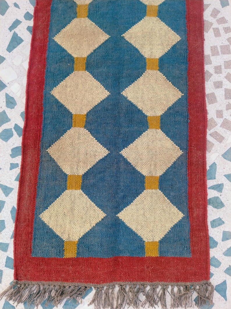 May include: A rectangular rug with a blue centre featuring a diamond pattern in cream and gold. The rug has a red border and a fringe at the bottom. The rug is made of woven material.