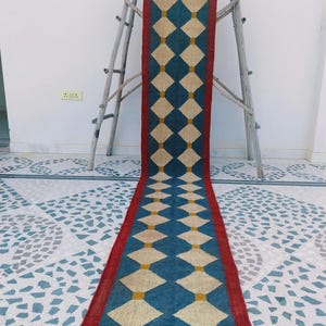 May include: A long, rectangular rug with a geometric diamond pattern. The rug features a blue background with cream-colored diamonds and yellow accents. It has a red border and gray fringe at the ends. The rug is displayed on a wooden ladder.