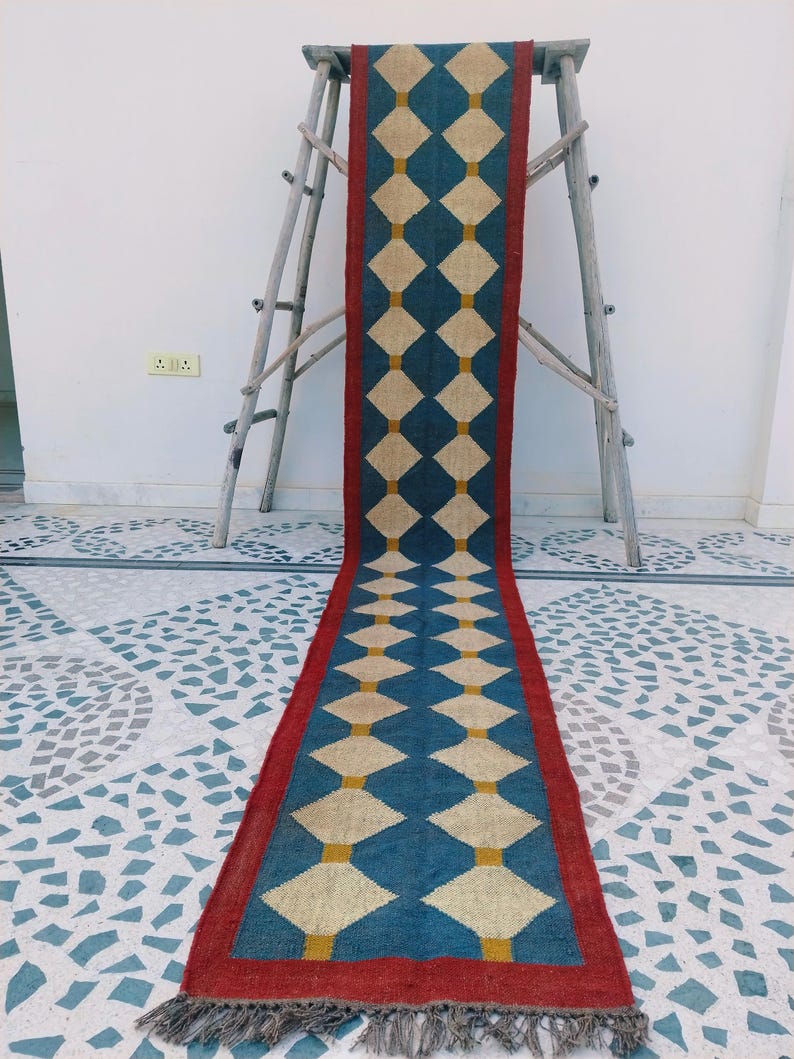 May include: A long, rectangular rug with a geometric diamond pattern. The rug features a blue background with cream-colored diamonds and yellow accents. It has a red border and gray fringe at the ends. The rug is displayed on a wooden ladder.