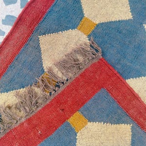 May include: A close-up of a woven rug with a geometric pattern. The rug features a blue background with cream-colored diamond shapes and yellow squares. A red border frames the design, and the rug has a fringed edge.