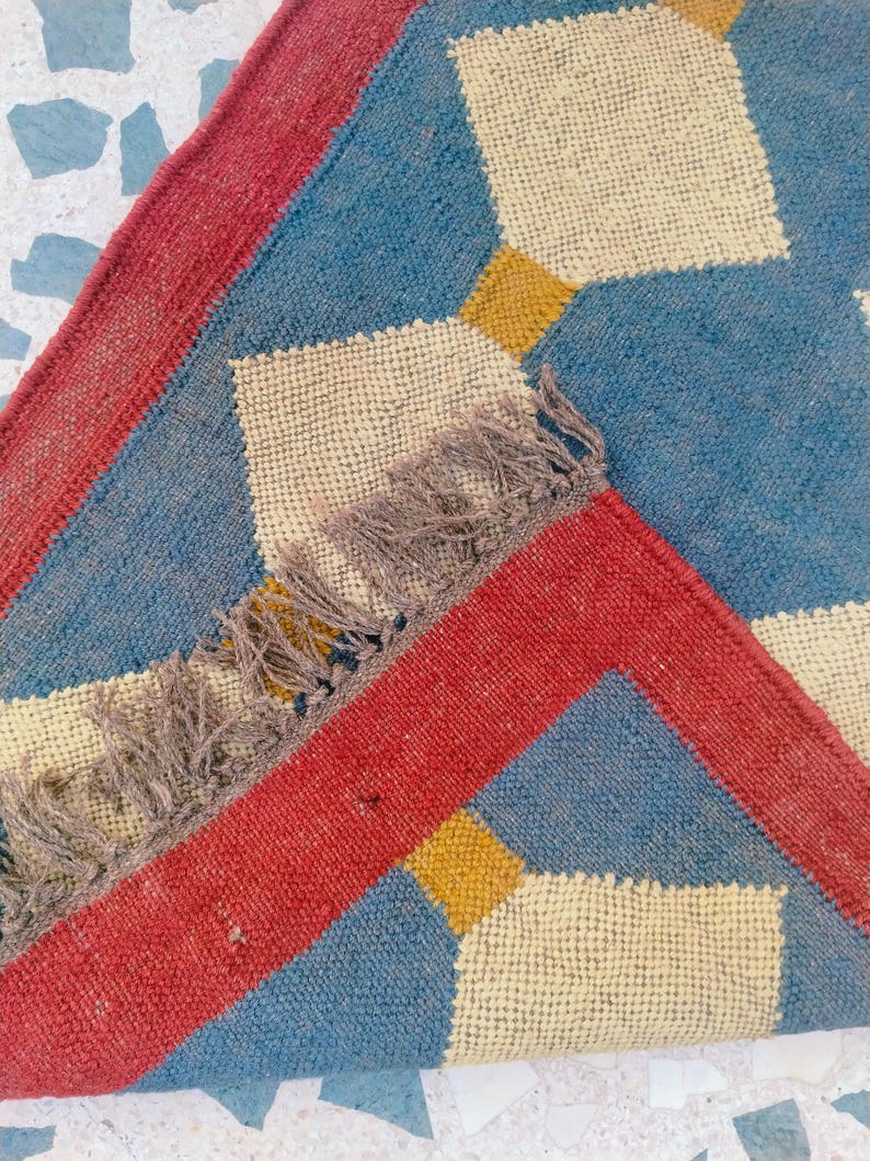 May include: A close-up of a woven rug with a geometric pattern. The rug features a blue background with cream-colored diamond shapes and yellow squares. A red border frames the design, and the rug has a fringed edge.