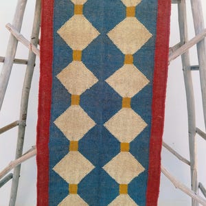 May include: A rectangular blue and beige patterned rug with a red border. The rug features a repeating diamond pattern in beige, connected by vertical gold lines. The rug is displayed on a wooden structure.