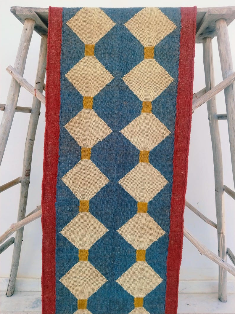 May include: A rectangular blue and beige patterned rug with a red border. The rug features a repeating diamond pattern in beige, connected by vertical gold lines. The rug is displayed on a wooden structure.