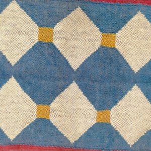 May include: Close-up of a woven rug with a repeating diamond pattern. The diamonds are white, set against a blue background, and connected by gold squares. The edges of the rug are red.