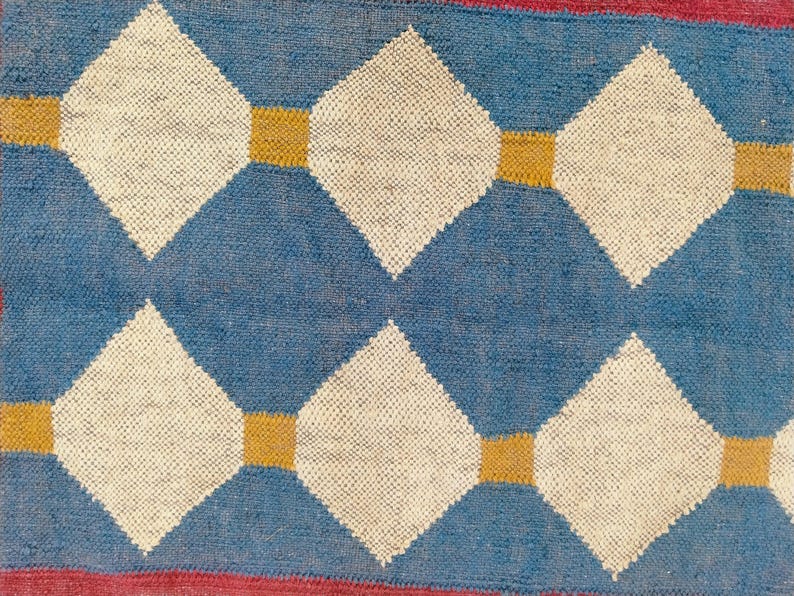 May include: Close-up of a woven rug with a repeating diamond pattern. The diamonds are white, set against a blue background, and connected by gold squares. The edges of the rug are red.