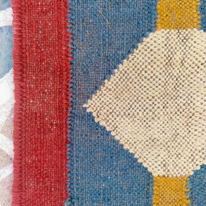 May include: Close-up of a woven textile with a geometric pattern. The design features a red border, blue sections, and off-white diamond shapes with yellow squares. The texture appears coarse and handmade, suggesting a rug or fabric.