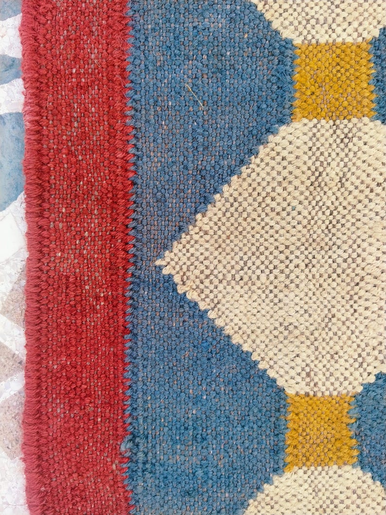 May include: Close-up of a woven textile with a geometric pattern. The design features a red border, blue sections, and off-white diamond shapes with yellow squares. The texture appears coarse and handmade, suggesting a rug or fabric.