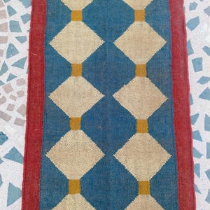 May include: A rectangular rug with a blue central field featuring a repeating pattern of cream-colored diamonds connected by gold rectangles. The rug is bordered by a red stripe on each side.