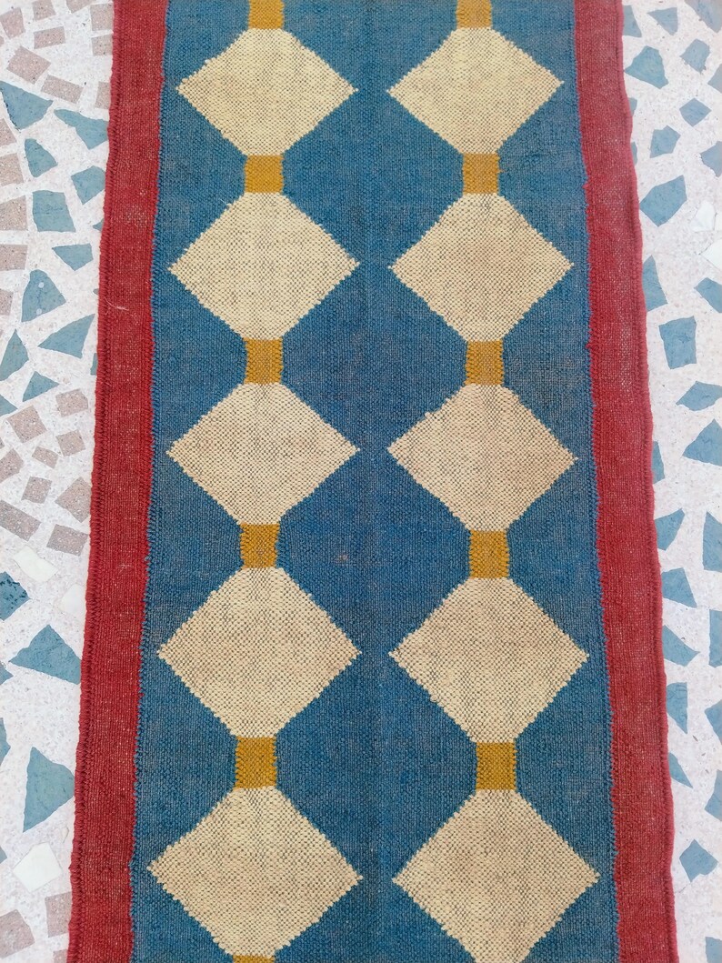 May include: A rectangular rug with a blue central field featuring a repeating pattern of cream-colored diamonds connected by gold rectangles. The rug is bordered by a red stripe on each side.