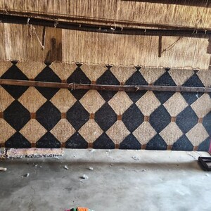 May include: A large, unfinished rug in a loom, featuring a repeating pattern of black and tan diamonds. The rug is framed by a wooden structure and supported by metal and wood components. The rug is in the process of being woven.