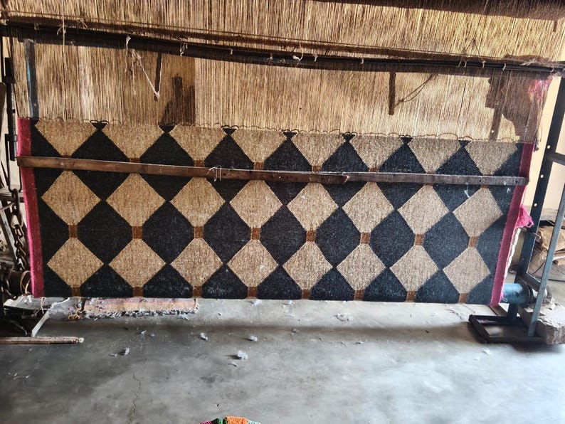May include: A large, unfinished rug in a loom, featuring a repeating pattern of black and tan diamonds. The rug is framed by a wooden structure and supported by metal and wood components. The rug is in the process of being woven.
