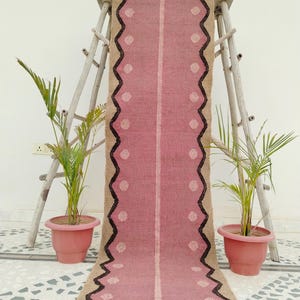 May include: A pink and tan runner rug with a central light stripe and dotted accents. The rug has a zigzag border in dark brown and is displayed on a wooden ladder. The rug is approximately 60 inches long.