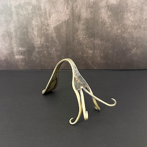 May include: A vintage, brass-toned fork bent into a decorative shape. The fork's tines are curved, and the handle is arched. The background is a dark surface with a textured gray wall.