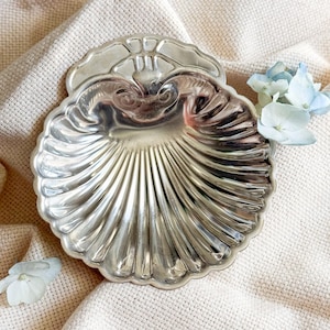 May include: A silver-colored, shell-shaped dish with a scalloped edge. The dish has a detailed, ridged interior and a decorative handle. Light blue flowers are placed near the dish on a cream-colored fabric.
