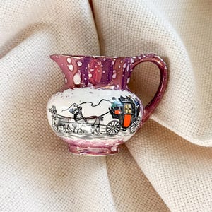 May include: A small, vintage ceramic creamer with a detailed illustration of a horse-drawn carriage. The creamer features a white body with a purple rim and handle, and a gold trim. The carriage scene is in black and white with touches of orange.