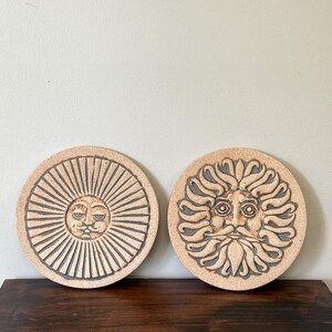 May include: Two circular ceramic wall plaques featuring relief designs. The left plaque displays a sun with straight rays and a central face. The right plaque shows a sun with wavy, flame-like rays and a detailed face with a beard and mustache.