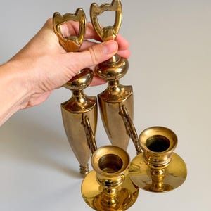 May include: A set of vintage brass candlesticks and bottle openers. The candlesticks have a round base and a cup for the candle. The bottle openers have a unique cat-shaped design. The items are gold in color.