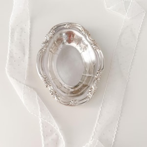 May include: An oval silver-toned metal dish with a decorative scalloped edge and embossed detailing, surrounded by a long, sheer white ribbon featuring a subtle dotted pattern. The dish has a polished, reflective surface and a deep center.