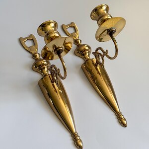 May include: Two vintage brass wall sconces. Each sconce features a candle holder with a circular drip plate, a decorative loop, and a long, pointed base. The sconces have a polished, golden finish and are designed to be mounted on a wall.