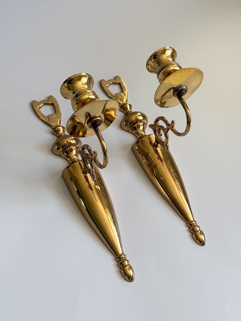 May include: Two vintage brass wall sconces. Each sconce features a candle holder with a circular drip plate, a decorative loop, and a long, pointed base. The sconces have a polished, golden finish and are designed to be mounted on a wall.